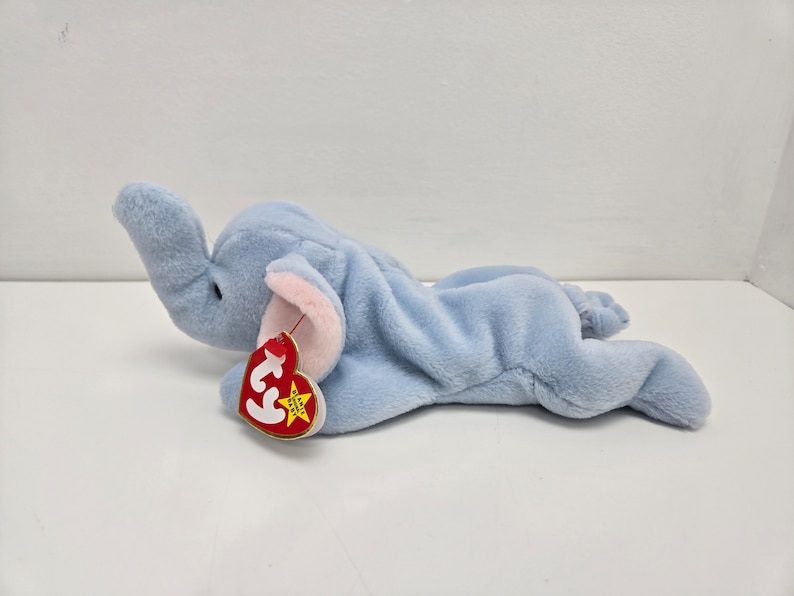 May include: A light blue plush elephant toy with a pink nose and ears. The toy is lying on its side with its head turned to the side. The toy has a red and yellow Ty tag attached to its leg.