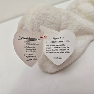 May include: A white plush toy named Fleece from The Beanie Babies Collection. The toy has a heart-shaped tag with the name "Fleece" and a poem about singing lullabies. The tag also includes the date of birth: March 21, 1996.