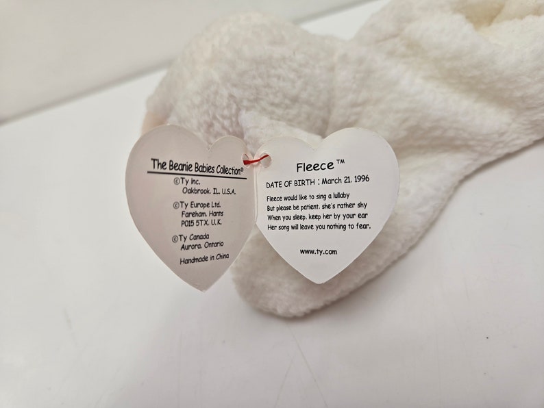 May include: A white plush toy named Fleece from The Beanie Babies Collection. The toy has a heart-shaped tag with the name "Fleece" and a poem about singing lullabies. The tag also includes the date of birth: March 21, 1996.