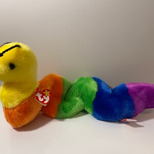 Op de afbeelding: Een gele en oranje beanie baby worm met een regenboogkleurig gestreept lichaam. De worm heeft een zwart gezicht met een kleine zwarte mond en zwarte ogen. De beanie baby heeft een rood Ty-label.