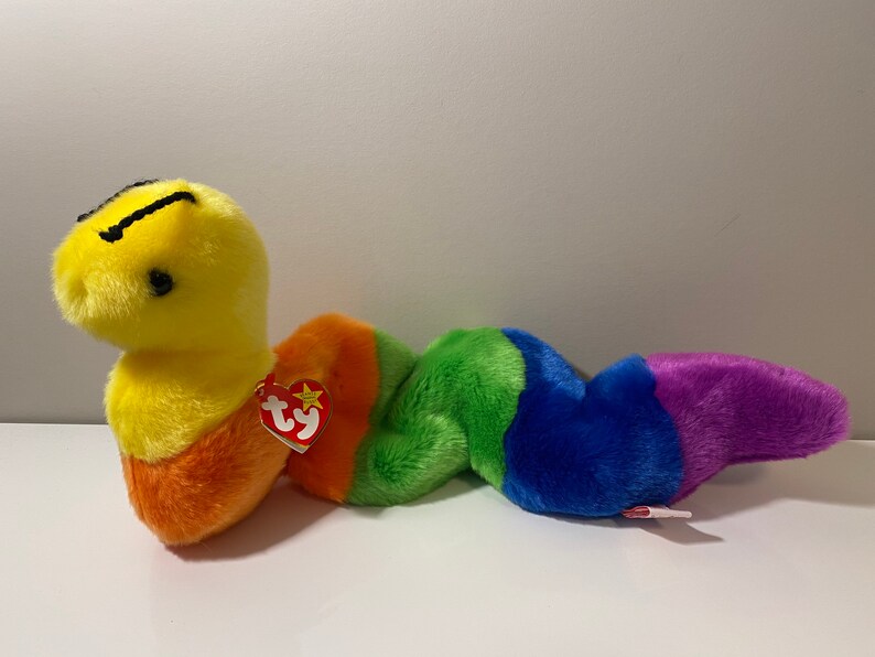 Op de afbeelding: Een gele en oranje beanie baby worm met een regenboogkleurig gestreept lichaam. De worm heeft een zwart gezicht met een kleine zwarte mond en zwarte ogen. De beanie baby heeft een rood Ty-label.