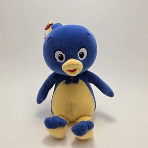 Peut inclure: Une peluche représentant un personnage avec une tête ronde bleue, de grands yeux blancs et un bec jaune. Le corps est bleu avec un ventre jaune, un petit nœud papillon bleu au cou et des pieds jaunes avec des orteils bleus. Une étiquette rouge et blanche est fixée au sommet.