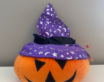 Puede incluir: Calabaza de felpa naranja con una cara sonriente negra y un sombrero de bruja morado con estrellas y lunas plateadas. El sombrero tiene un borde negro.