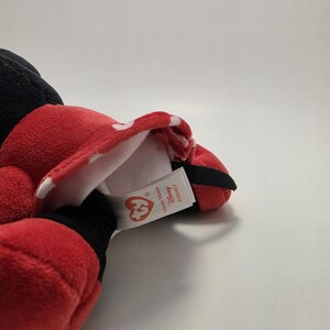 May include: A red plush Minnie Mouse doll with a white polka dot bow and black accents. The doll has a tag that reads "Ty" and "Disney". The doll is lying on a white surface.