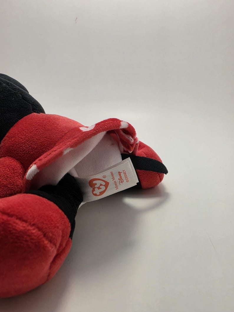 May include: A red plush Minnie Mouse doll with a white polka dot bow and black accents. The doll has a tag that reads "Ty" and "Disney". The doll is lying on a white surface.