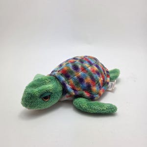 May include: A soft toy turtle with a green head, flippers, and tail. The shell is made of a patchwork of colourful squares in red, blue, yellow, and purple. This children's toy is designed to be cuddled.