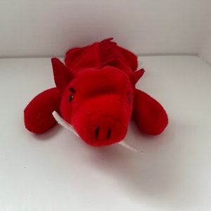 May include: Red plush dragon toy with white teeth and black eyes. The dragon is lying on its belly with its head facing the camera.