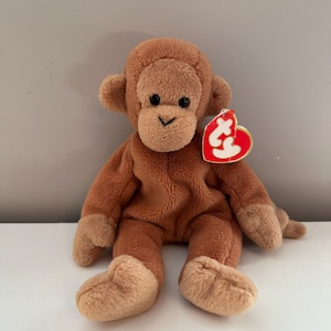 May include: A brown plush monkey toy with a red heart-shaped tag that says "Ty" on it. The monkey is sitting with its legs crossed and has a brown nose and black eyes.