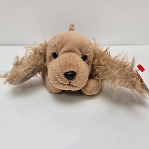 May include: A brown and tan plush toy dog with floppy ears. The dog has a red tag attached to its ear.