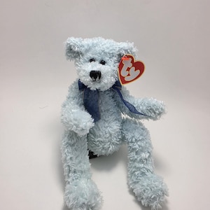Può includere: Un orsacchiotto di peluche azzurro con pelliccia riccia, occhi in plastica nera e naso cucito nero. L'orso indossa un nastro blu scuro legato al collo e presenta un'etichetta Ty rossa a forma di cuore attaccata all'orecchio sinistro con le lettere ty.