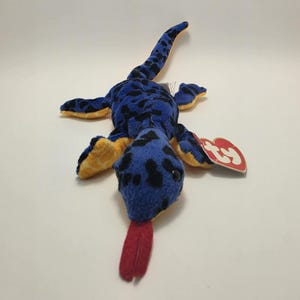 Puede incluir: Un juguete de lagarto de peluche con cuerpo azul con manchas negras y parte inferior de color amarillo brillante. El animal de peluche tiene una lengua larga de fieltro rojo, ojos de plástico negro y una etiqueta Ty roja en forma de corazón.