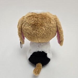 May include: A brown and white plush dog with pink ears sitting on a white background. The dog has a black spot on its back.