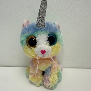 Puede incluir: Un juguete de peluche de gato unicornio colorido con un cuerno plateado brillante, ojos grandes azules y una nariz rosa. El gato está sentado con las piernas cruzadas y tiene un patrón de pelaje de arcoíris.