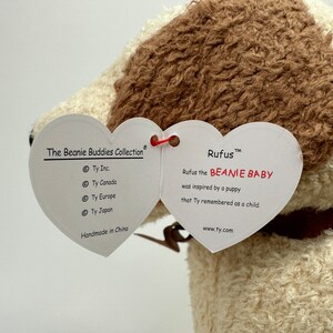 Op de afbeelding: Een wit en bruin Beanie Baby pluche speelgoed genaamd "Rufus" met een hartvormig label. Het label luidt "Rufus the BEANIE BABY was inspired by a puppy that Ty remembered as a child." Het label bevat ook het Ty Inc. logo en de website www.ty.com.