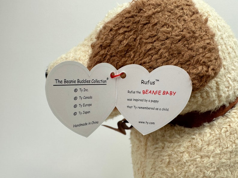 Op de afbeelding: Een wit en bruin Beanie Baby pluche speelgoed genaamd "Rufus" met een hartvormig label. Het label luidt "Rufus the BEANIE BABY was inspired by a puppy that Ty remembered as a child." Het label bevat ook het Ty Inc. logo en de website www.ty.com.