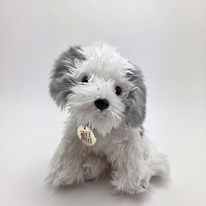 May include: A soft toy dog with white and grey fur, seated on a white surface. The toy has a small tag with Chinese characters. The dog's fur is shaggy, and its ears and head are grey. The toy is a cute, cuddly design.