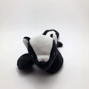 May include: A plush toy badger in black and white, lying on a white surface. The badger has a white face with black markings and small black eyes. Its body is black, and its paws are visible.