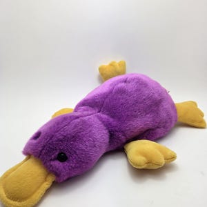 May include: A purple plush platypus toy with yellow feet and bill. The toy has a soft, fuzzy texture and black eyes. The platypus is lying down on a white surface. The toy is suitable for children.