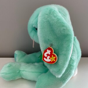 Puede incluir: Un peluche de conejo verde menta con orejas largas y ca&iacute;das. El peluche tiene una etiqueta roja en forma de coraz&oacute;n sujeta a su oreja. La etiqueta tiene las palabras "BEANIE BUDDY" y un logotipo.