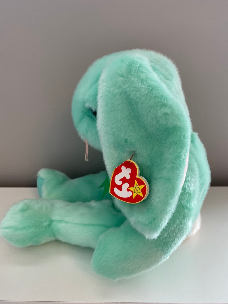 Puede incluir: Un peluche de conejo verde menta con orejas largas y ca&iacute;das. El peluche tiene una etiqueta roja en forma de coraz&oacute;n sujeta a su oreja. La etiqueta tiene las palabras "BEANIE BUDDY" y un logotipo.