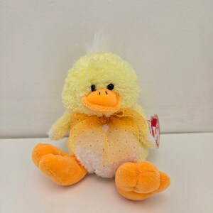 May include: A plush yellow duck toy with an orange beak and feet. The duck has a fluffy body, a sheer orange bow with white polka dots, and a white belly. A red tag is attached to the duck.