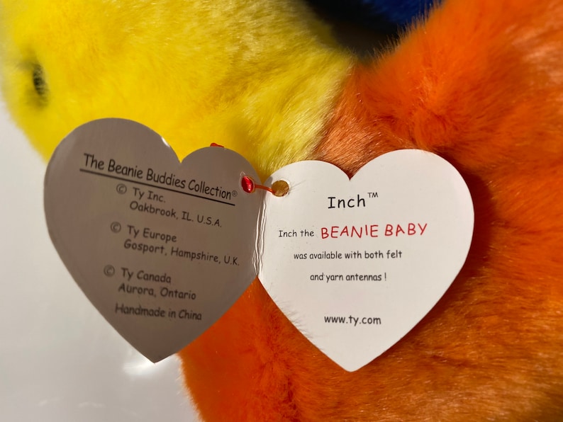 Op de afbeelding: Een gele, oranje en blauwe Beanie Baby genaamd Inch met een hartvormig label. Het label luidt "Inch the Beanie Baby was available with both felt and yarn antennas!" en "www.ty.com".