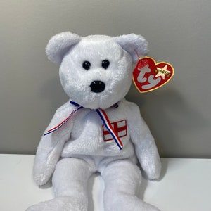 May include: White plush beanie baby bear wearing a red, white, and blue ribbon with an embroidered English flag on the chest. The bear has black eyes and a black nose. The bear is sitting on a white surface. The bear has a red Ty Beanie Babies tag attached to its right ear.