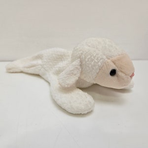 May include: A plush white lamb toy with a soft, textured body. The lamb has a light pink face, black eyes, and a small pink nose. The toy is lying down, with its legs extended.