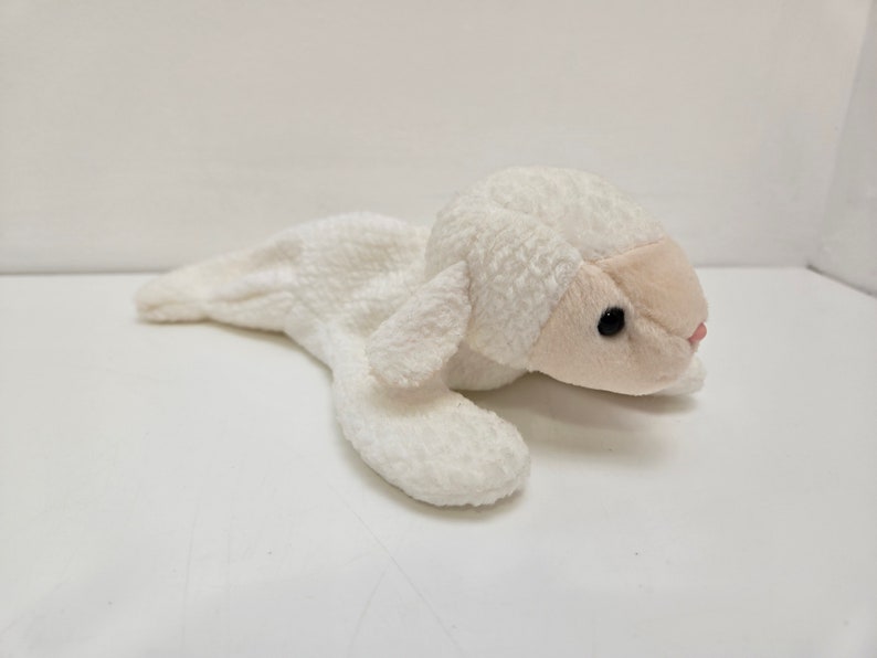May include: A plush white lamb toy with a soft, textured body. The lamb has a light pink face, black eyes, and a small pink nose. The toy is lying down, with its legs extended.