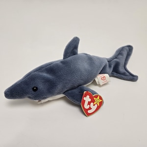 May include: A gray plush shark toy with a white belly and a red Ty tag that says "Beanie Baby" with a star.
