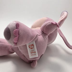 K&ouml;nnte beinhalten: Ein rosa-lila Pl&uuml;schtier mit gro&szlig;en Ohren und einem unverwechselbaren Gesicht. Das Spielzeug hat ein Etikett mit der Aufschrift "Angel Disney". Das Pl&uuml;schtier liegt auf der Seite und zeigt seinen weichen, strukturierten Stoff.