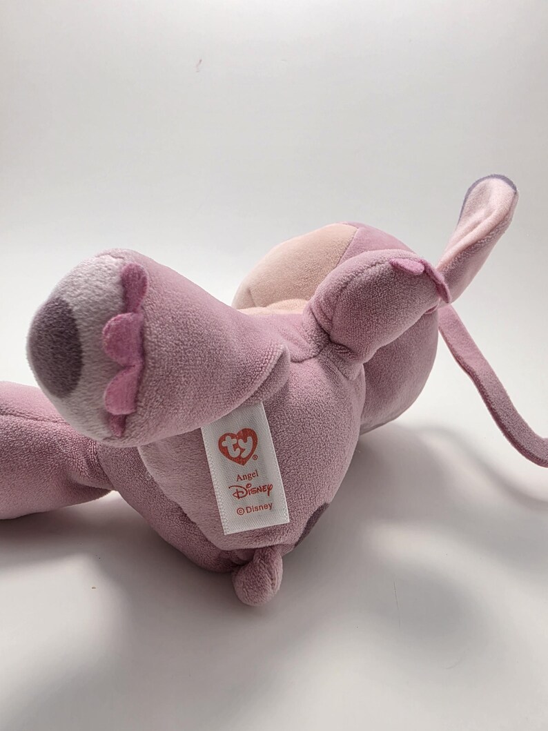 K&ouml;nnte beinhalten: Ein rosa-lila Pl&uuml;schtier mit gro&szlig;en Ohren und einem unverwechselbaren Gesicht. Das Spielzeug hat ein Etikett mit der Aufschrift "Angel Disney". Das Pl&uuml;schtier liegt auf der Seite und zeigt seinen weichen, strukturierten Stoff.