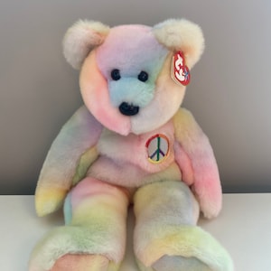 May include: A soft, rainbow-coloured teddy bear with a peace sign patch on its chest. The bear has a Ty tag attached to its ear. The bear is made of fluffy material and has black eyes and nose.