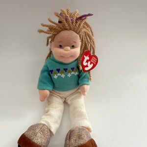 Può includere: Una bambola di peluche con lunghi capelli biondi, che indossa un maglione blu con un motivo floreale, pantaloni bianchi e scarpe marroni. La bambola ha un fiocco viola nei capelli e un'etichetta rossa Ty che dice "Ty Inc. Beanie Babies".