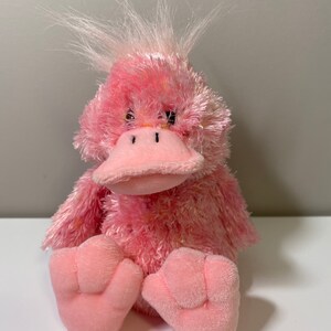 May include: Pink plush duck with a fluffy white crest. The duck has a pink beak and black eyes.
