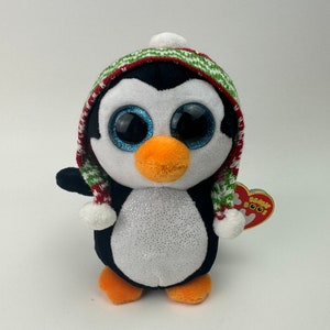 May include: A white and black penguin plush toy wearing a red, green, and white striped hat with a white pom-pom. The penguin has large blue eyes and a glittery white belly. The toy has a Ty Beanie Boo's tag attached.