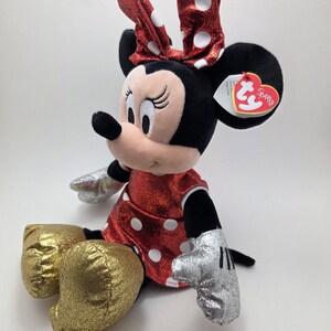 May include: A plush Minnie Mouse doll. She has a red sparkly dress and bow, both with white polka dots. The doll has black ears, a pink face, gold glittery shoes and silver gloves. A Ty heart-shaped tag is attached.