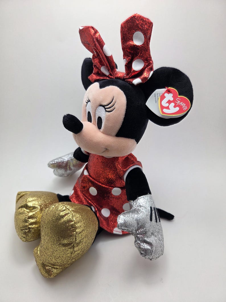 May include: A plush Minnie Mouse doll. She has a red sparkly dress and bow, both with white polka dots. The doll has black ears, a pink face, gold glittery shoes and silver gloves. A Ty heart-shaped tag is attached.