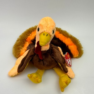 May include: A brown and orange plush turkey with a yellow beak and feet. The turkey has a red wattle and a Ty tag attached to its wing.