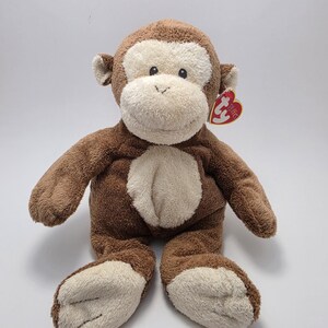 May include: A plush monkey toy with brown fur and a cream-colored face, belly, and feet. The stuffed animal has a red heart-shaped tag attached to its ear. The monkey is sitting with its arms and legs outstretched.