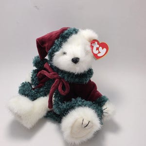Può includere: Un orsacchiotto di peluche bianco che indossa un cappello di velluto rosso scuro e un cappotto di velluto rosso scuro abbinato con finiture in pelliccia sintetica verde scuro. L'orso ha un naso nero e un'etichetta Ty rossa e gialla a forma di cuore.