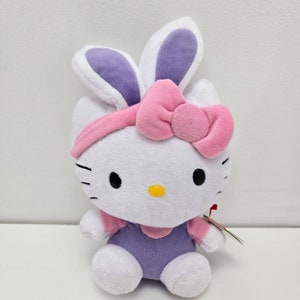 以下が含まれることがあります： 黒い目とひげを持つ白いぬいぐるみ猫。ピンクのリボンと紫色の内側の耳が付いたウサギの耳を着用しています。猫はピンクの袖が付いた紫色の衣装を着ています。小さなタグが付いています。