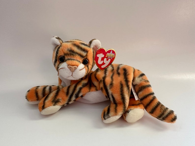 Op de afbeelding: Een pluche tijger knuffeldier met oranje en zwarte strepen, witte poten en een witte buik. De tijger ligt op zijn buik met zijn kop naar de zijkant gedraaid. Hij heeft een rood Ty hartvormig label met het woord "Ty" en het jaar "2000" erop gedrukt.