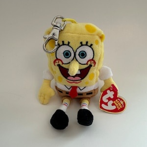 May include: A yellow SpongeBob SquarePants plush toy with embroidered eyes and a red tie. The plush has a metal clip and a Ty Beanie Babies tag attached. The toy is approximately 4 inches tall.