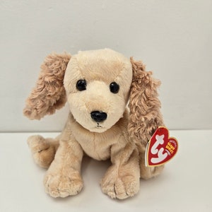May include: A plush golden retriever dog toy with floppy ears and a black nose. The stuffed animal has large black eyes and a red heart-shaped tag attached to its ear. The toy is sitting on a white surface.