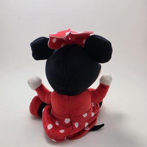 May include: A plush Minnie Mouse doll, viewed from the back, with a black head and ears, a red dress with white polka dots, and a matching bow. The doll's arms are outstretched, and the background is plain white.