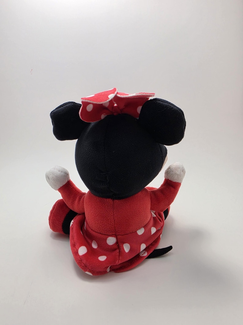 May include: A plush Minnie Mouse doll, viewed from the back, with a black head and ears, a red dress with white polka dots, and a matching bow. The doll's arms are outstretched, and the background is plain white.