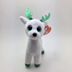 May include: A white plush reindeer toy with green antlers and accents. The reindeer has large, round eyes with green irises and a black nose. A red and yellow tag is attached to the ear.