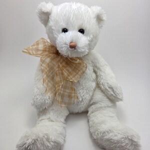 May include: A white teddy bear with a light brown checked bow. The bear has black eyes and a pink nose. The soft toy is sitting with its legs extended.