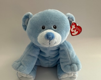 Può includere: Un orsacchiotto di peluche blu chiaro con dettagli bianchi sulle zampe e sul naso. L'orso è seduto con le zampe davanti a sé. L'orso ha un'etichetta Ty rossa con la parola "Puffy" su di essa.